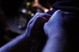 Close up of two hands in fists placed upon someone's back. The image is dark and barely lit. In the background there is an out of focus figure seated in a wheelchair, but barely visible. 