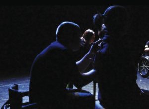 Dark and silhouetted image of a person seated in a wheelchair with their hands on an audience member's back. Audience member is seated on a stool. Both people are in profile and dressed in black. In the background is another pair in the same relationship. 