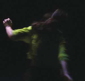 Blurry and dark, close-up image of dancer mid-movement taken from behind. She's wearing a neon yellow t-shirt and has a long ponytail. The details are out of focus.